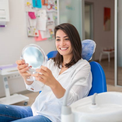 Young Woman Looking At Mirror With Smile In Dentist’s Office Sealants